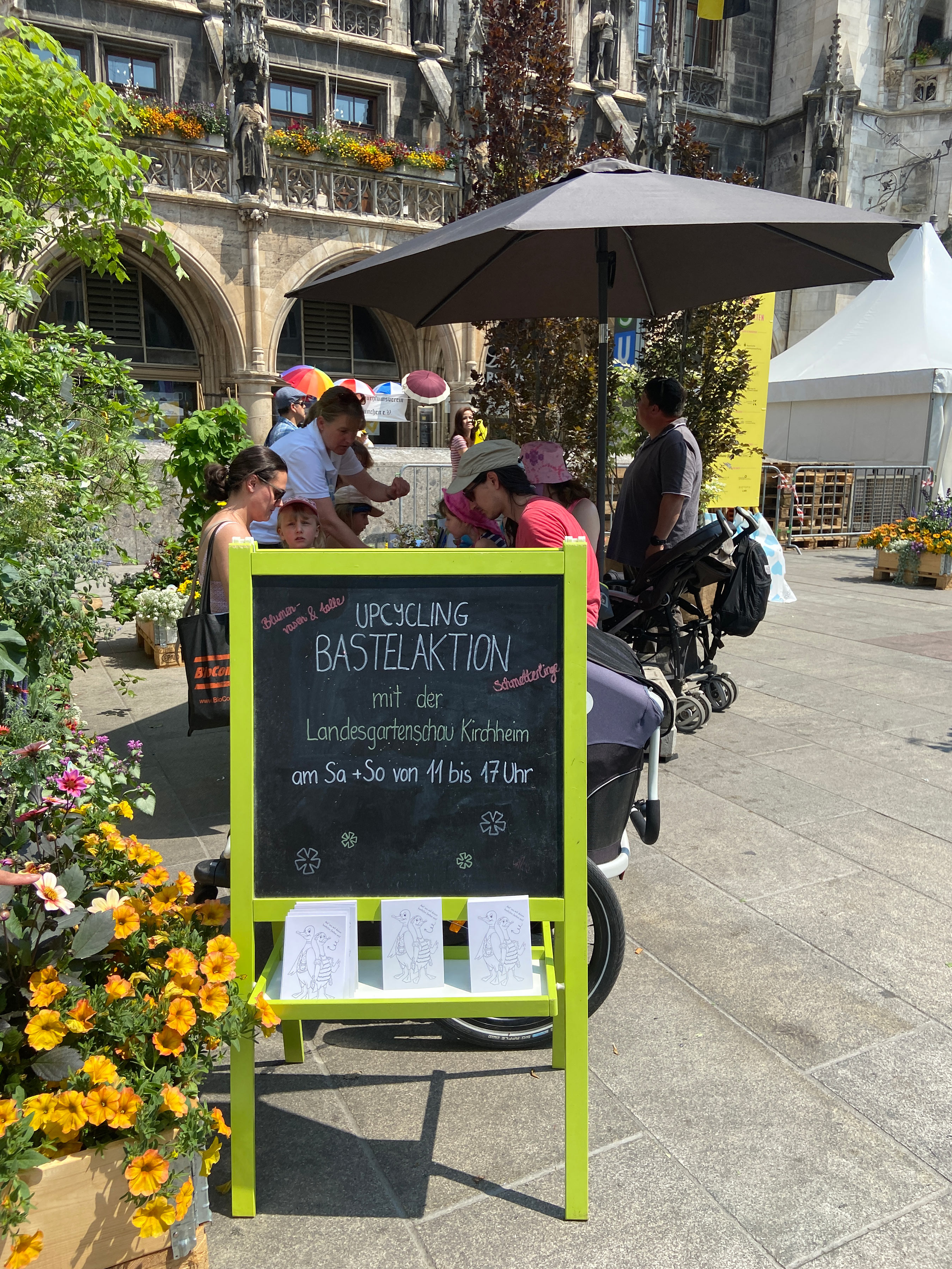 Im Vordergrund Schiefer-Aufstelltafel Upcycling Bastelaktion 
Im Hintergrund Kinder und Erwachsene unter einem Sonnenschirm und das Münchner Rathaus